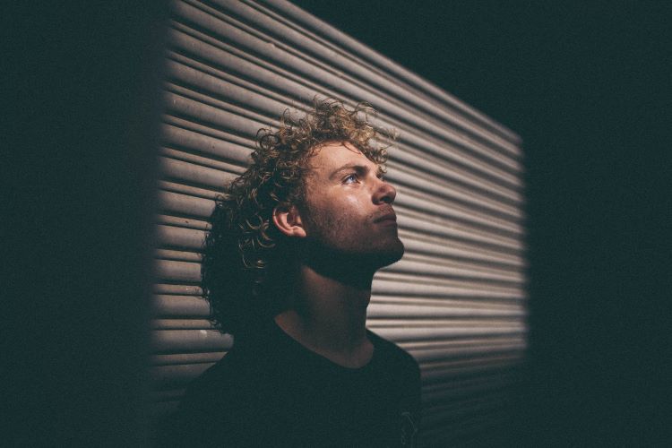 A man in darkness with his back against a shutter, looking up