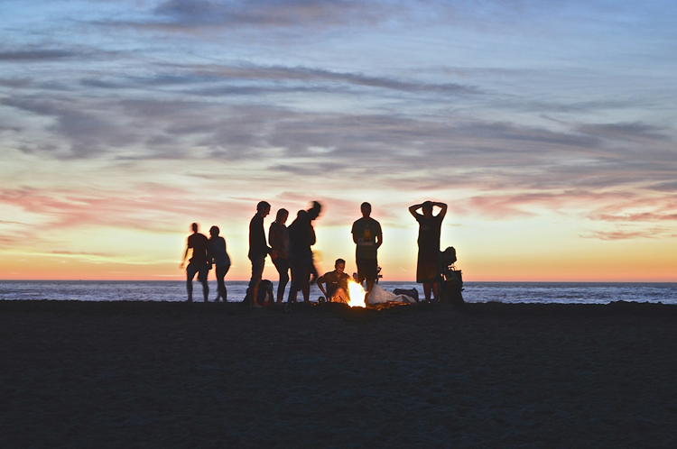 A group of people in silhouette, around a fire at the beach during sunset